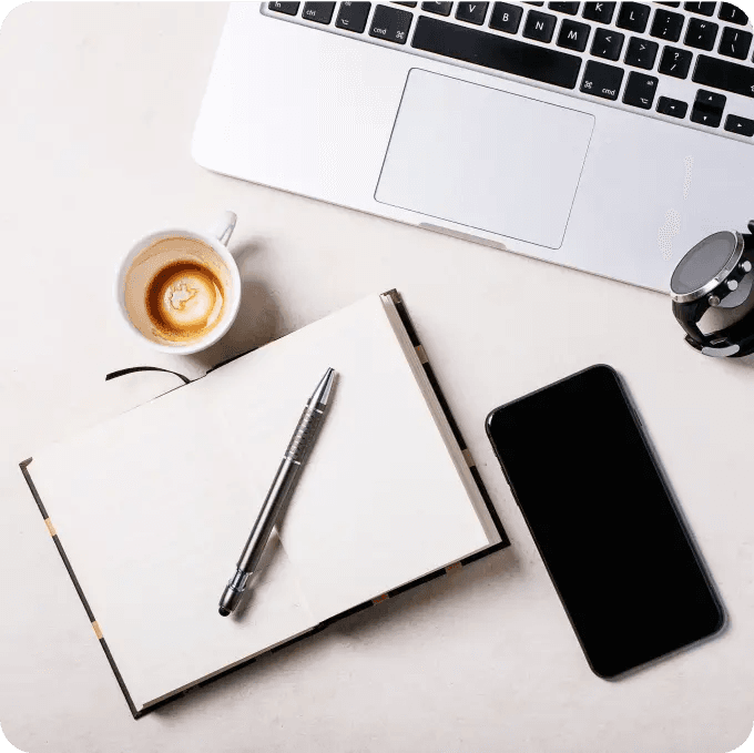 Biurko, na którym znajduje się notes, srebrny długopis, smartfon, laptop, czarny zegarek ze srebrną tarczą i kawa w białym kubku.