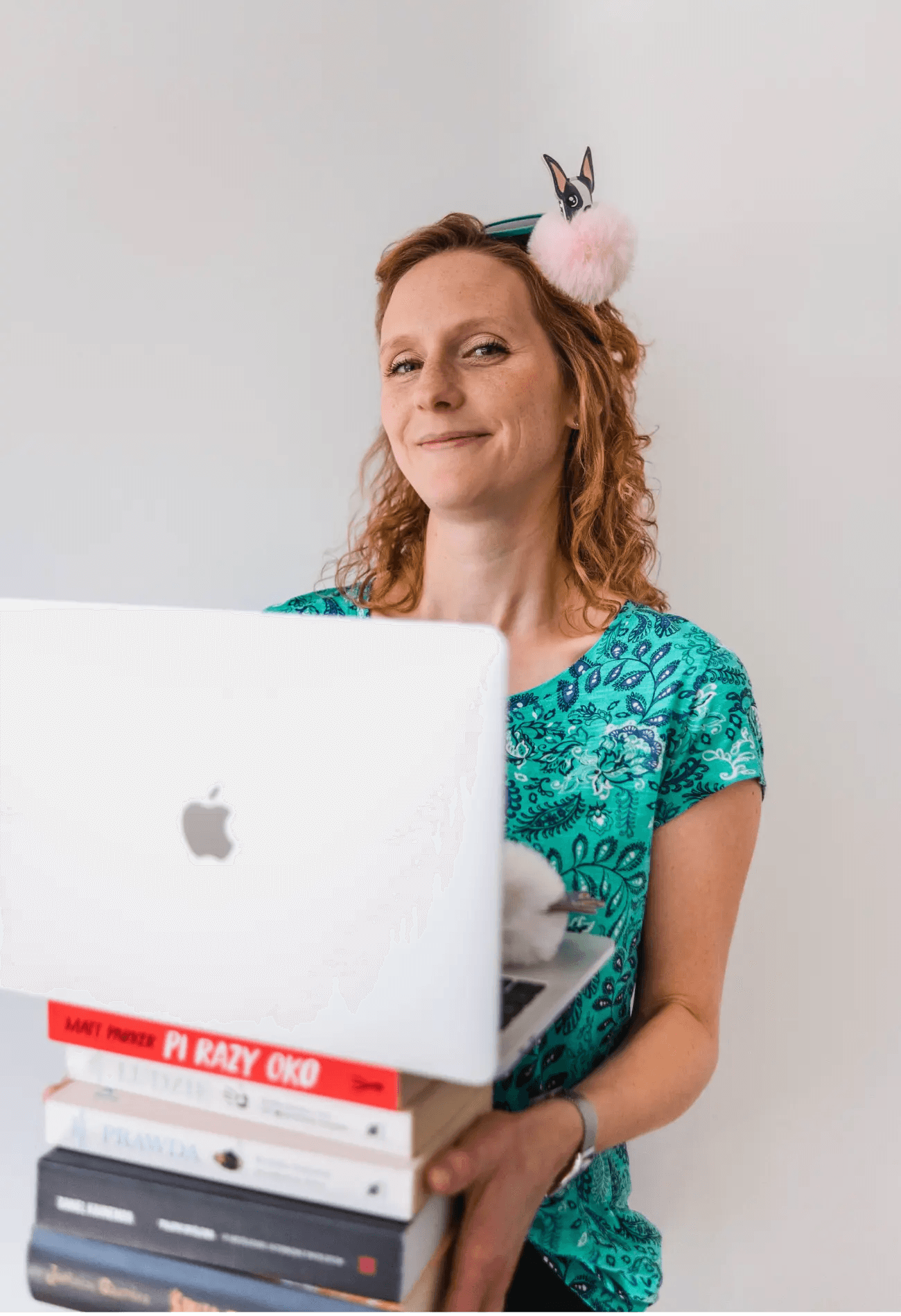 Uśmiechnięta kobieta w zielonej wzorzystej bluzce, w dłoniach trzyma książki oraz laptopa.