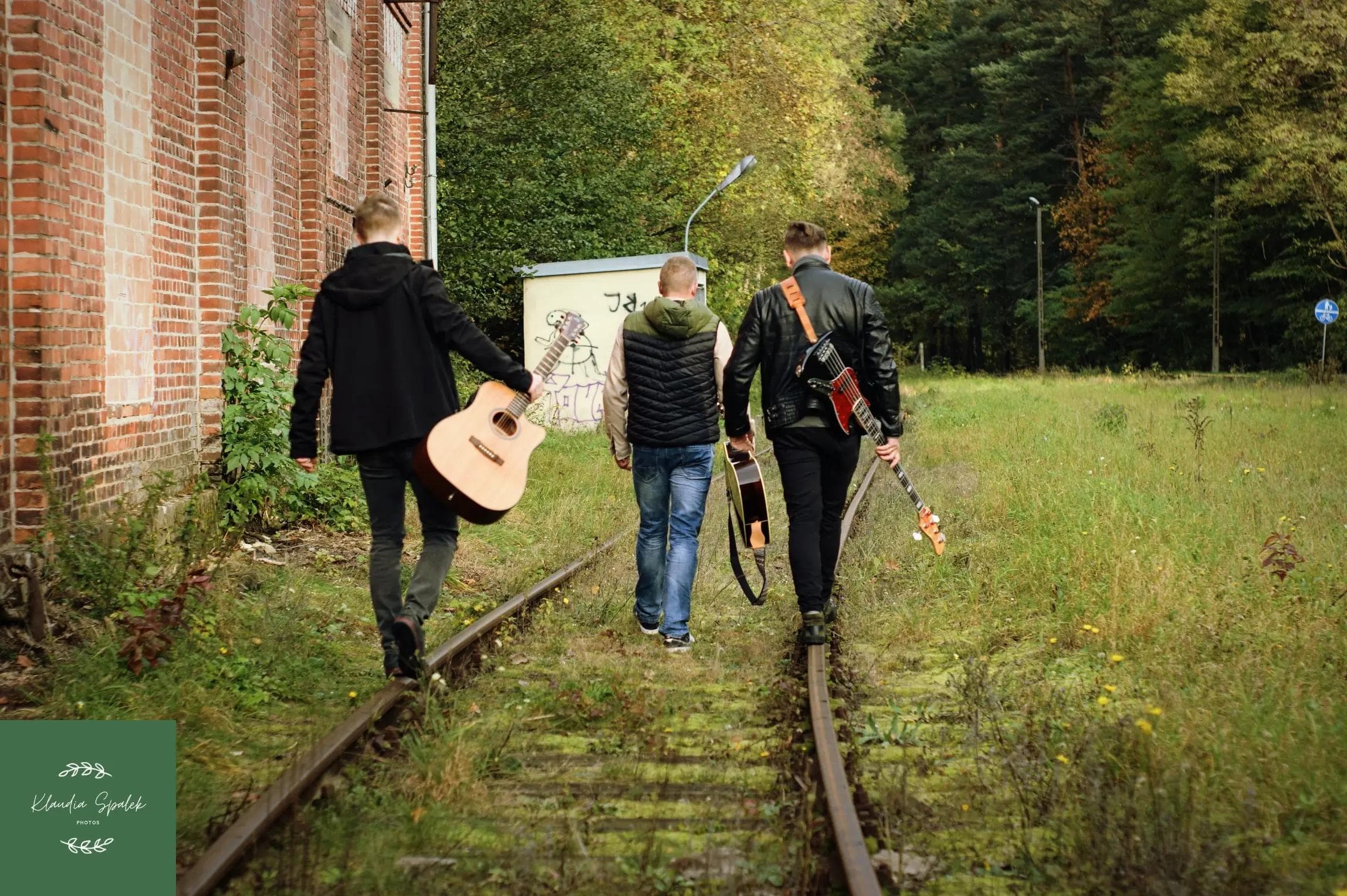 Trzech mężczyzn z zespołu Kumple Bejdaka z gitarami w dłoniach, idą po torach kolejowych odwróceni tyłem, w tle widać drzewa, po prawej ceglany budynek.